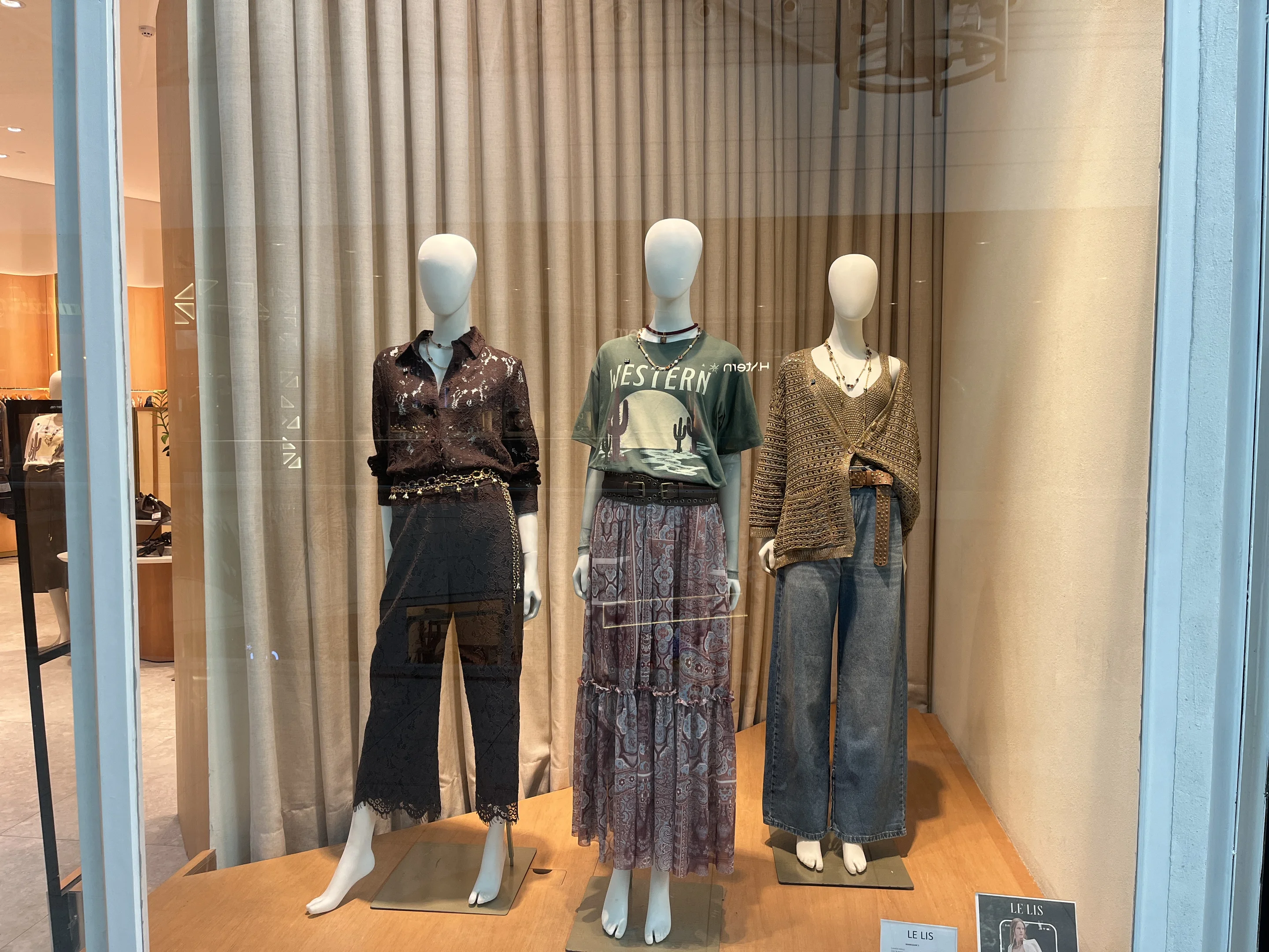 Foto da vitrine da loja Lelis com 3 manequins femininos. Todos estão vestindo roupas na paleta de cores entre o marrom e vinho. As peças são da coleção outono-inverno.