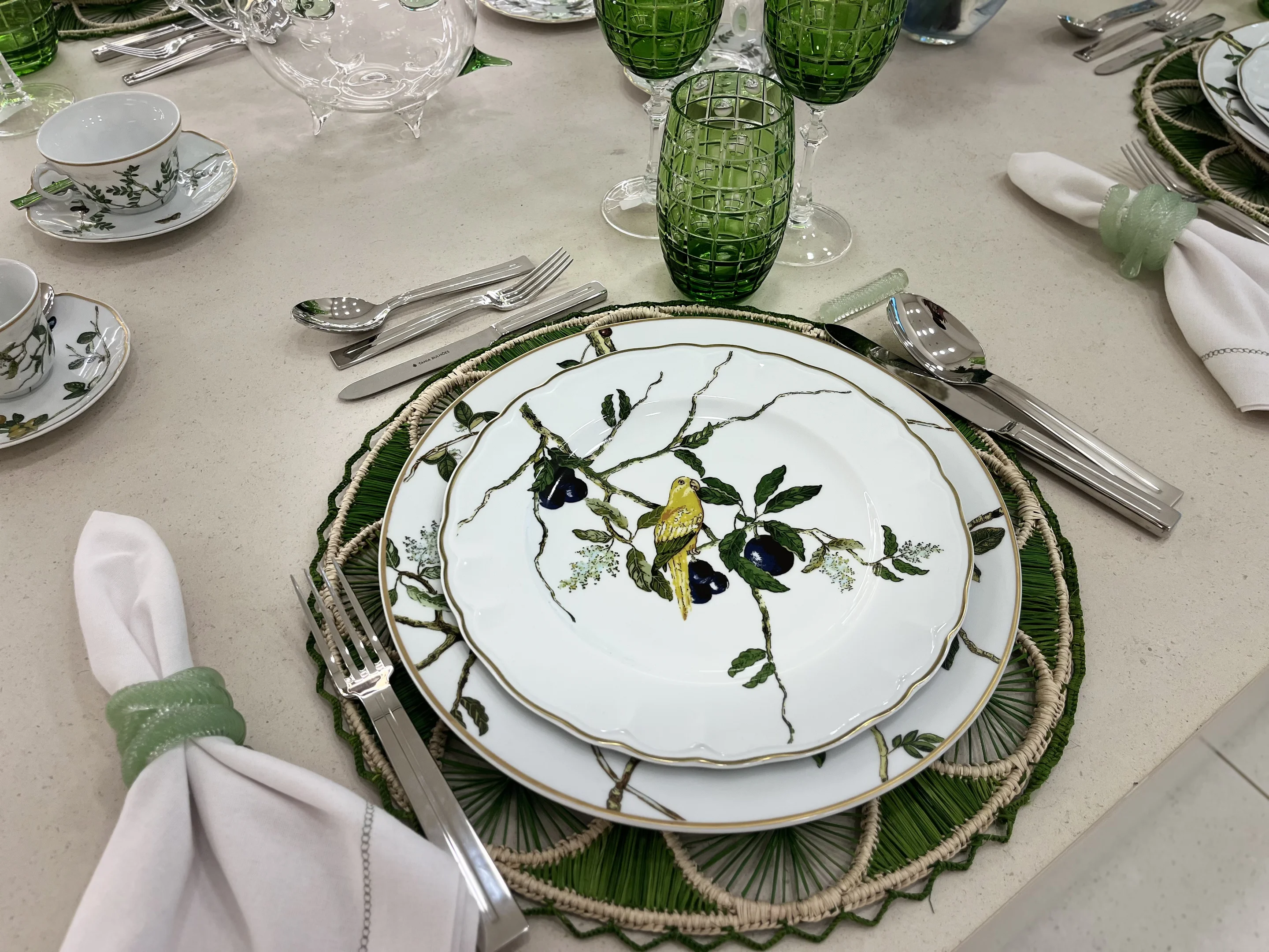 Mesa posta com itens em tons verdes, prato com um passarinho pintado de amarelo e algumas folhas verdes em galho de árvore.