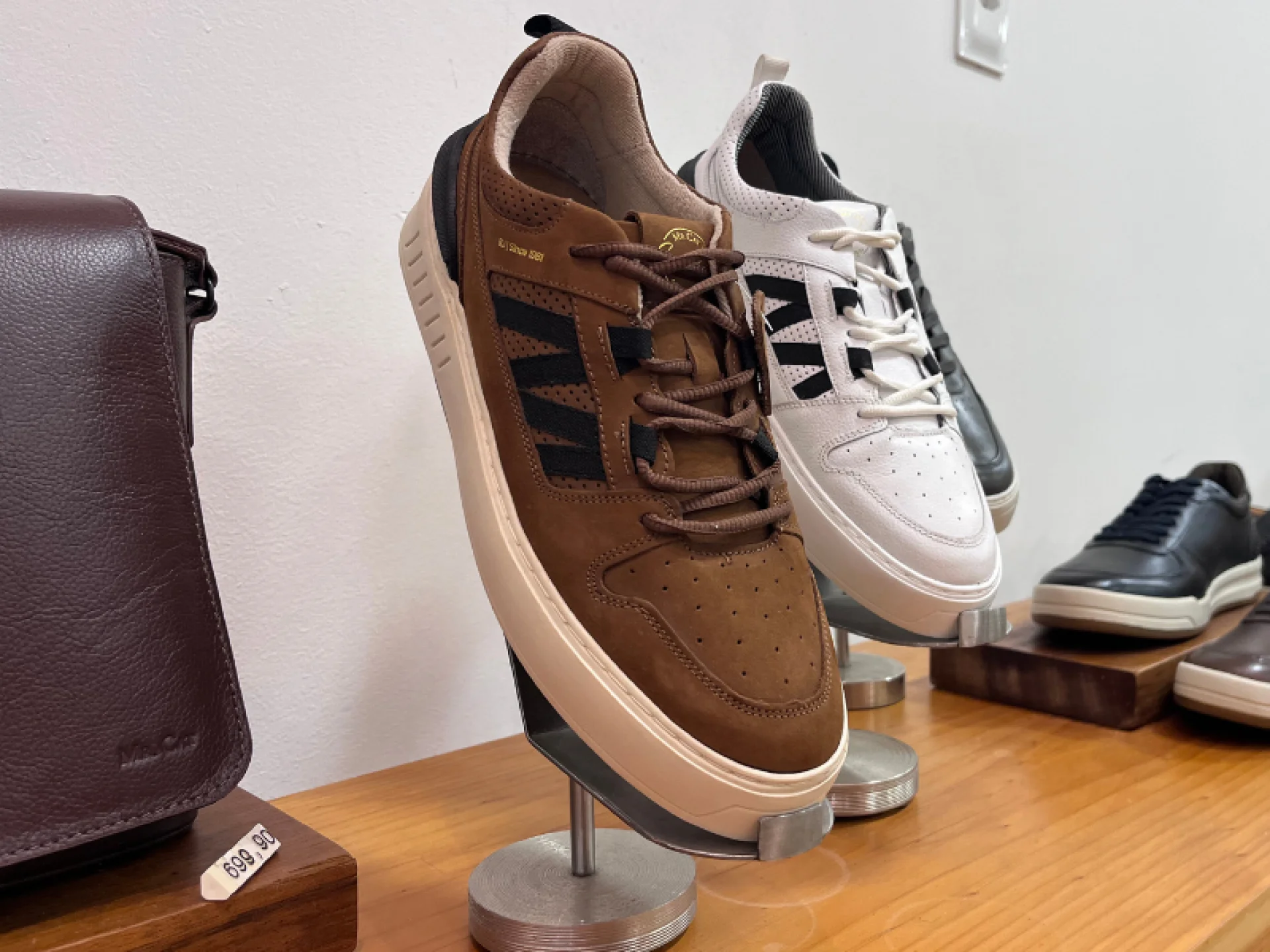 Tênis em exposição da cor marrom com detalhes preto na lateral e base branca. Ao fundo, tênis do mesmo modelo da cor branca.